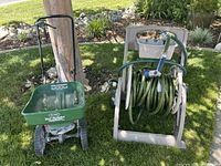Side view of the Scotts green lawn spreader next to the beige hose reel with green garden hose coiled on it and attached hose nozzle.