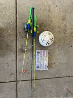 Image showing two children's fishing rods with reels and yellow fishing lines attached to bobbers, a clear plastic tackle box containing fishing lures, and a white soccer ball with black patterns all laid out on a concrete floor.