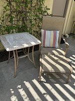Photo of outdoor beige lounge chair with armrests, multicolor striped pillow, and mosaic tile top square side table in sunlight next to a plant.