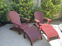 Two teak Adirondack chairs with matching ottomans on concrete patio with greenery background.