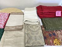 Grouped stack of various rectangle tablecloths showing solid red, green, beige, a heart pattern, and other intricate designs in mixed materials.