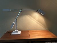 Desk lamp illuminated, showing the arm extended over a wooden surface, casting light on a textured wall.
