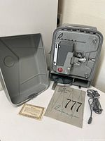 Photo showing the Revere 777 projector standing next to its open protective carrying case. The original manual and power cord are placed on the surface in front of the projector.