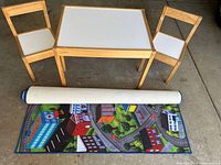 Photo showing the wooden children's table and two chairs with white surfaces, alongside the rolled city-themed play mat.