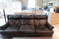 Full view of black/dark brown leather 3-seater sofa in a living room and kitchen setting.