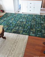 Blue and green geometric patterned area rug shown covering part of a light carpet and darker wood flooring near white furniture.