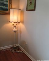 Full view of the floor lamp with white scalloped lampshade lit and standing on wooden floor near wall.