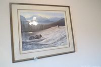 Framed limited edition print showing mountain landscape with bear and wolf in snow. Front view with window light reflections.