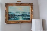 Front view of framed oil painting showing ocean waves and ornate gold frame hanging on a wall next to a lamp.
