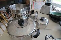 Set of four Lagostina stainless steel pots including steamer/double boiler stacked and arranged on kitchen counter with other kitchen items around.