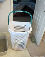 Front full view of the tall white plastic laundry basket with ventilation holes and a light blue handle, positioned on floor near a door and a litter box.
