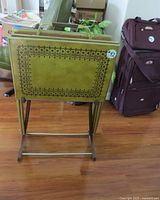 Four stacked vintage green metal TV trays with decorative dark border pattern shown from the side on the storage stand with wheels.