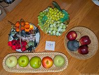 Photo showing wooden apples in long wicker tray, wooden apples and pear in small wicker basket, yellow glass grapes on plate with handle, and clusters of plastic grapes in red, amber, and pink on serving dish.
