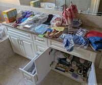 Countertop with various bathroom-related items including bathing accessories, heating pads, curlers, dusters, and open cabinet with various toiletries and items inside.