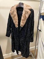 Full front view of black textured coat with brown fur collar hanging on a hanger against a beige wall.