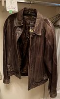 Front view of brown Danier leather jacket hanging on hanger showing zippered front, elastic cuffs, and collar snap button. Visible Thinsulate lining label inside jacket.