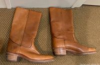 Pair of men's Frye leather boots laid side by side on carpet, showing full view of outer sides, stacked heels, and toe shape. Brown leather with visible wear and creases.