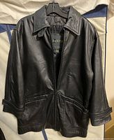 Front view of black leather jacket on hanger, showing collar, zipper, and pockets.