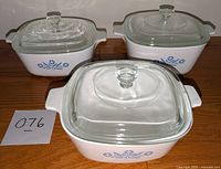 Three Corning Ware casseroles with glass lids arranged on a wooden surface.