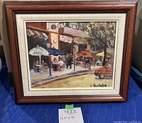 Full frontal photo of the framed oil painting featuring a street cafe scene with tables, umbrellas, a red car and trees, with artist signature visible