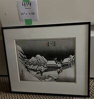 Full view of framed black and white Japanese wood-block print art, showing a snowy village landscape with mountains and figures carrying loads, framed in black frame with white mat.