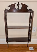 Front view of wooden hanging shelf showing three horizontal shelves, dark wood finish, decorative carved top with finial, and criss-cross side supports.