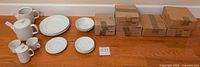 Photo of Johnson Brothers Athena dishes lot staged on wooden floor showing cups, mugs, saucers, bowls, plates, coffee pots, and creamer with several unopened boxes to the right.