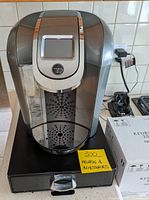 Front angle shot of the Keurig 2.0 coffee machine showing the digital screen, water reservoir, and drip landing area.