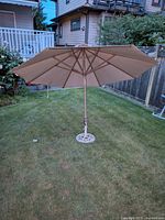 Full view of large beige outdoor umbrella opened and angled, mounted on metal stand placed on grass lawn with background houses and fences.