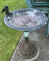 Side angle showing full bird bath with cherub figure on rim, weathered bowl and ornate base on patio next to green plastic chairs and grass