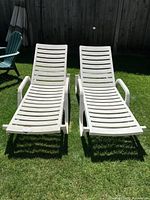 Two white plastic slatted outdoor lounge chairs with armrests photographed side by side on a grass lawn.