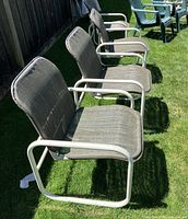 Three vintage Samsonite patio chairs on grass outdoors, showing white tubular metal frames and greenish-gray woven mesh fabric seating and backs.