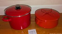 Two red pots with lids on wooden floor, one tall with glass lid, one shorter with red lid.