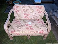 Loveseat with floral seat and back cushions arranged on brown wicker frame outdoors on grass.