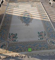 Large woven area rug laid flat outdoors under partial sun and shade. Shows full rug with floral border and central medallion design, fringed edges visible.