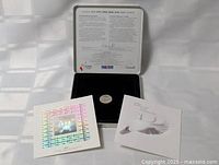 Opened metal tin box with coin, stamp, and certificate inside on white background with natural light.