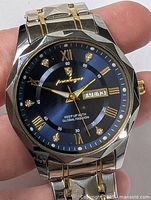 Close-up of the watch face showing the blue dial with gold hands, Roman numerals, day and date window, and brand logo.