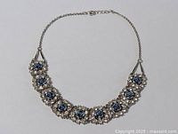 Full view of the vintage floral blue stone bracelet laid flat showing floral clusters with blue rose stones and clear rhinestones, metal chain with clasp and extender.