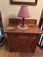 Front view of antique wooden cabinet with table lamp on top, showing drawer and two cupboard doors with brass handles and knob.