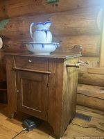 Side angle of antique wooden wash cabinet with ceramic water jug and wash basin on top, showing drawer and door details