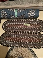 Photo showing several braided oval rugs including a rolled rug with navy and beige colors and two flat braided oval rugs in brown and black tones with dimension tags.