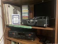 Photo of shelf containing vinyl records, cassette tapes stacked vertically, portable turntable in carrying case, and Denon double cassette player below