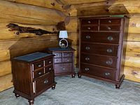 Photo showing two matching wooden nightstands and one tall wooden dresser in a log cabin style room, with a table lamp on one nightstand.
