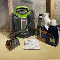 Bissell Little Green ProHeat spot remover with hose and four cleaning solution bottles placed on a wooden surface, alongside user manual.