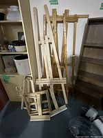 Four wooden easels grouped together against a wall. Three large easels are standing upright with vertical supports and adjustable canvas holders. One smaller easel rests in front, showing a simpler design.