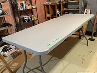 Full view of rectangular plastic top folding table with folded legs extended and sturdy metal frame, situated in a basement workshop setting.