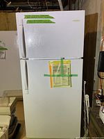 Front view of white Frigidaire refrigerator with top freezer, labels taped to doors, set in basement.