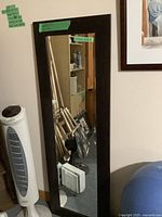 Brown framed rectangular mirror leaning against a wall, next to a tower fan. Mirror has a wide dark brown wooden frame and reflective mirror glass.