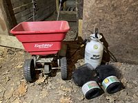 Wide view showing the EarthWay spreader, white Wood Mason pump sprayer, and three rolls of black garden edging mesh on the ground in a wood and dirt environment.