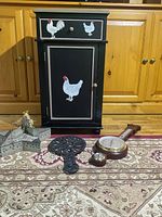Jam cupboard painted black with rooster and chicken designs on drawer and door, with iron trivet, star-shaped tray, and barometer placed on carpet in front of wooden cabinet wall.